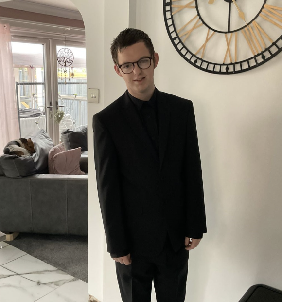 Zack, 19, is in a black suit and tie, looking excited to attend his prom. he has short hair, is white, and slim and wears glasses