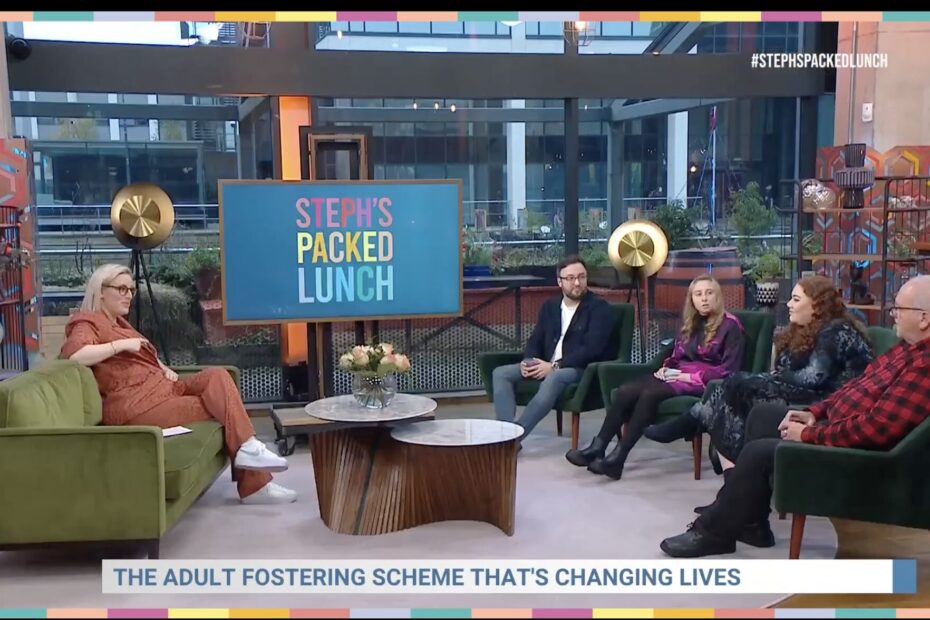 A freeze frame of pss' appearance on Steph's Packed Lunch. Steph sits on a coach to the left and the family sit on the right with a coffee table in between them and Steph. At the bottom of the screen reads a headline: adult fostering scheme changing lives