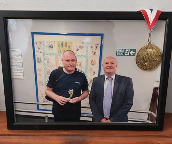 A photo frame displayed in the home. In the photo frame is a picture of James accepting a medal from his local day centre.