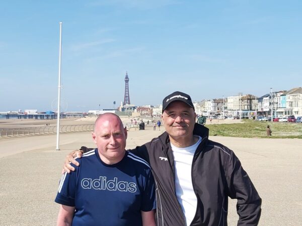 James and Adrian on holiday in Blackpool on a blue sky day. Adrian and James stand side by side with Adrian's arm around James' shoulder.