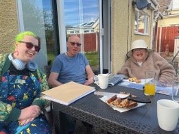 Hope has green hair and wonderful smile - she is sitting with two people we support in Shared Lives and having a nice slice of cake that they welcomed her with before they had a chat about their quality of support in PSS.