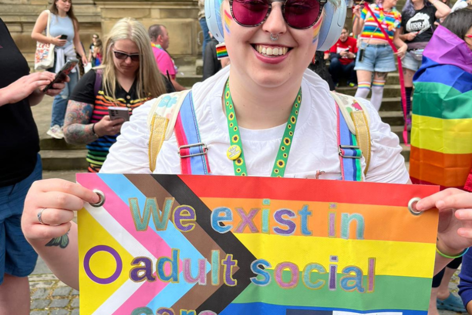 Hope has blue hair and wears purple tinted glasses. She has a huge smile and wears a white t shirt and multicoloured dungarees. She's holding up a handheld sign that has the LGBTQIA+ flag and reads 'we exist in adult social care'