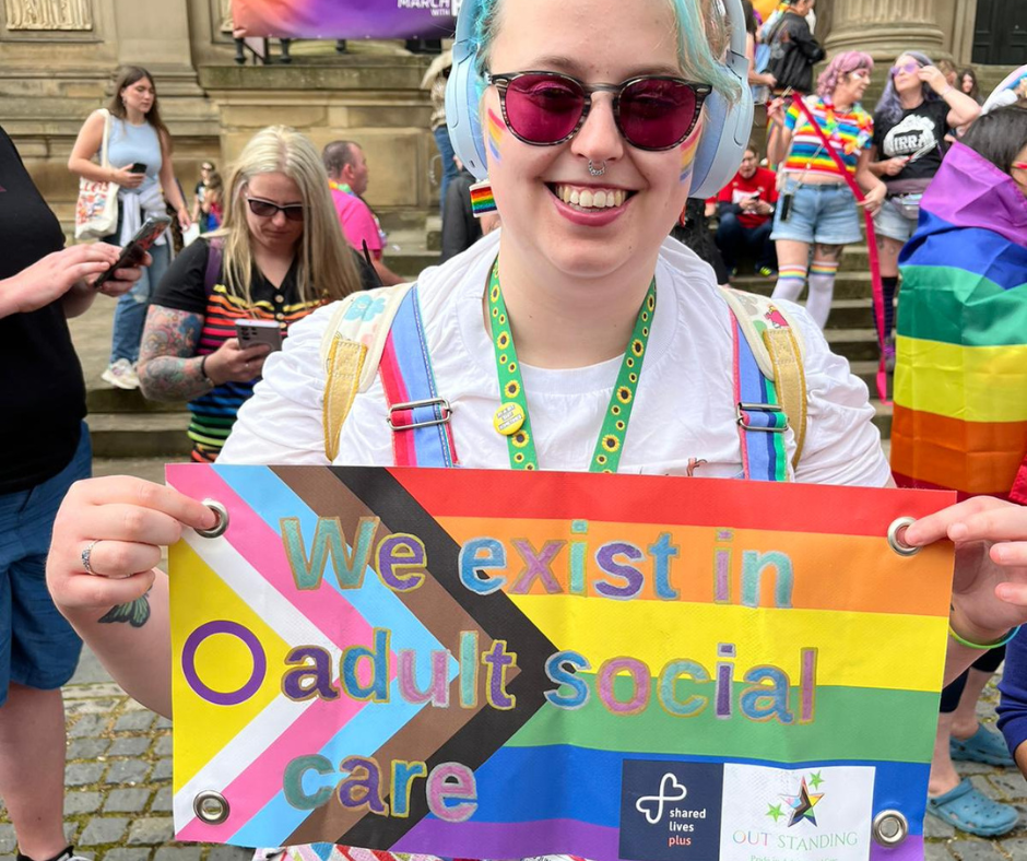 Hope has blue hair and wears purple tinted glasses. She has a huge smile and wears a white t shirt and multicoloured dungarees. She's holding up a handheld sign that has the LGBTQIA+ flag and reads 'we exist in adult social care'