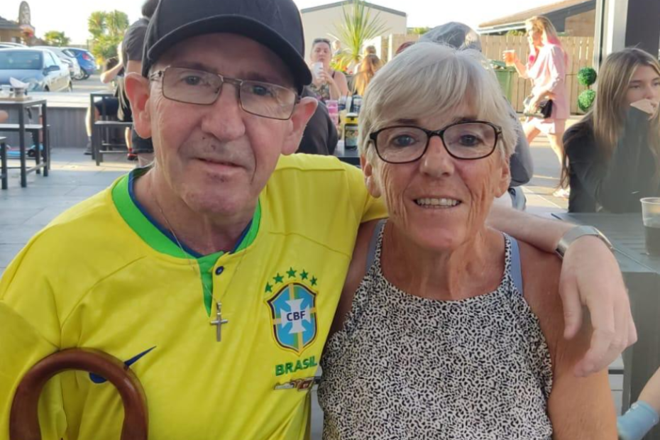 Kevin and Pauline stand close together, Kevin's arm around big sis, Pauline. They're close in age, they look around 50s/60s - Kevin and Pauline are white and both wear glasses. Kevin wears a yellow football shirt and black cap - his walking stick is just in shot. Pauline, wears a summery top and has short blonde/gray hair. they both looks really comfy with each other.