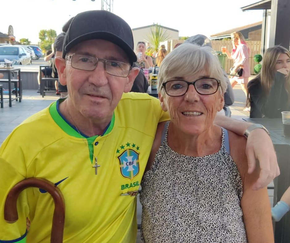 Kevin and Pauline stand close together, Kevin's arm around big sis, Pauline. They're close in age, they look around 50s/60s - Kevin and Pauline are white and both wear glasses. Kevin wears a yellow football shirt and black cap - his walking stick is just in shot. Pauline, wears a summery top and has short blonde/gray hair. they both looks really comfy with each other.