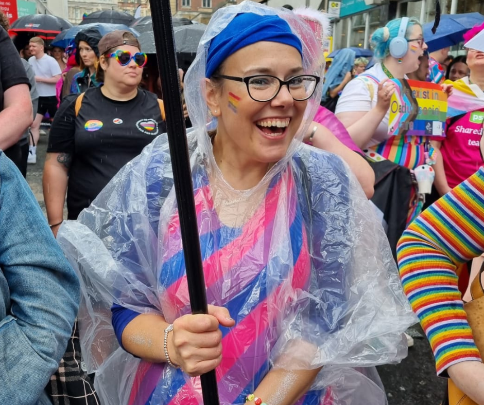 Mandy is smiling and has hold of the pole from the PSS Pride banner. She's wearing a dress with the bisexual flag colours and wears glasses. She has a blue head scarf on and is amongst a crowd of people.