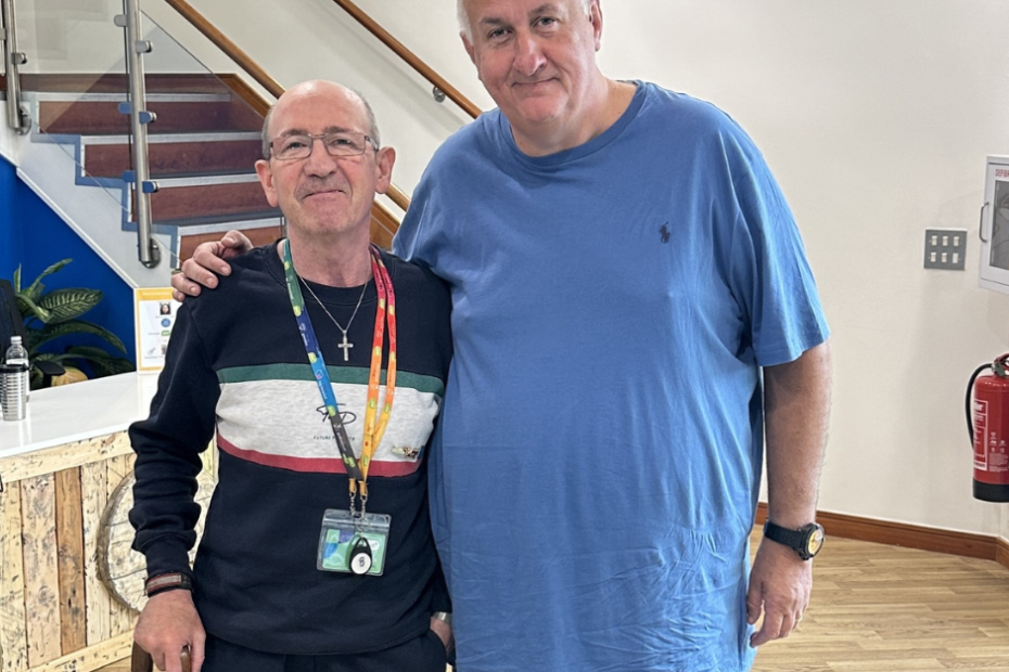 Paul and kevin stand together in the reception area of PSS. Paul is a tall man! He's head and shoulders above Kevin who is older than Paul and uses a walking stick. They're stood together like old friends, there's a lot of love and respect between them. Paul is white and has great short hair, he wears a baggy blue t shirt and jeans while Kevin as a PSS lanyard on because he's a volunteer at the offices.