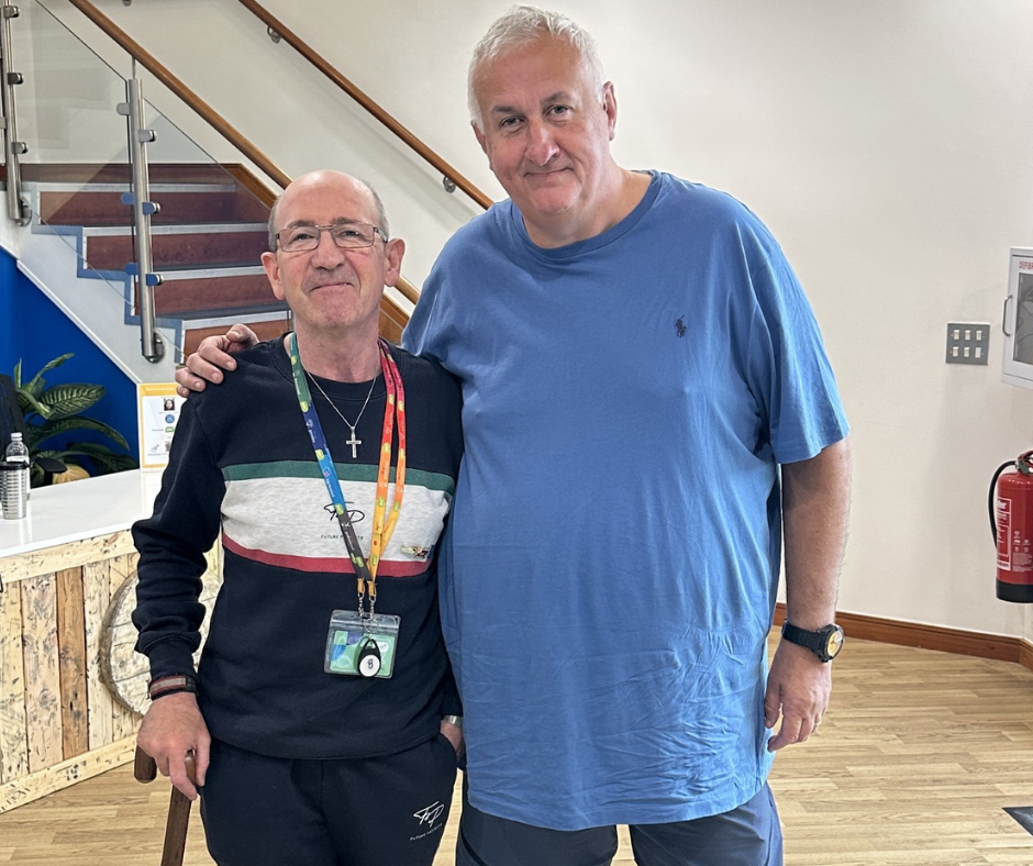 Paul and kevin stand together in the reception area of PSS. Paul is a tall man! He's head and shoulders above Kevin who is older than Paul and uses a walking stick. They're stood together like old friends, there's a lot of love and respect between them. Paul is white and has great short hair, he wears a baggy blue t shirt and jeans while Kevin as a PSS lanyard on because he's a volunteer at the offices.