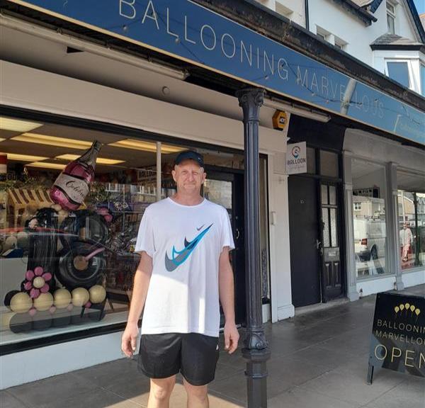 Paul is a white man, tall and slim. He's wearing a black cap, nike tshirt and black shorts - it's a lovely warm day in Llandudno. He's stood smiling outside the party shop that his Shared Lives carers own.