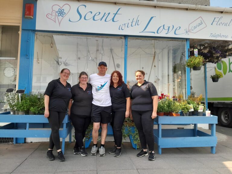 Paul takes a group photo with the women who work at the neighbouring store, Scent with Love. He has his arms wrapped around the shoulders of two and the other two stand either side. They look like good friends and happy to have Paul around. They're in front of the store front.