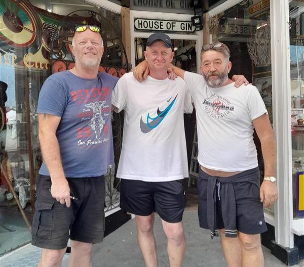 Paul stands in between two gents who run the antiques shop called The Treasure Chest. They are all wearing sports t shirts and shorts and looking like friends.