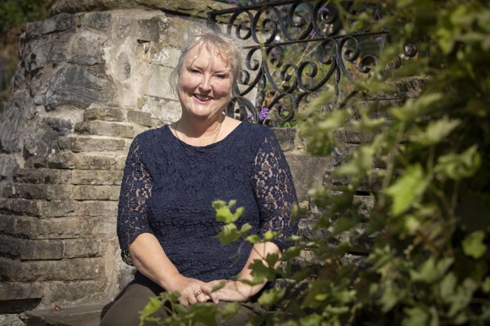 Karen sits in her garden on a sunny day - she smiles warmly at the camera and her hands are placed politely on her lap as she sits down among the trees. She's wearing a navy top and she's a blong, white woman in her 50s.