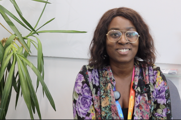 Lisa Maynard-Atem from Black United Representation Network sits in front of a white wall and next to a green houseplant. she is smiling and wearinga floral top and wide rimmed glasses. She is a Black woman with shoulder length brown hair