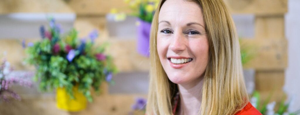 Simone is white, blonde and smile brightly, She's at the PSS office which has a pallett wall behind her with some colourful florals attached to it