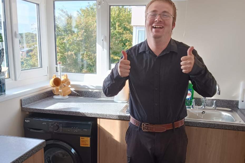 Andrew is a white man with ginger hair. He's standing in his new kitchen with two thumbs up and a beaming smile. He's so excited to be in his own place