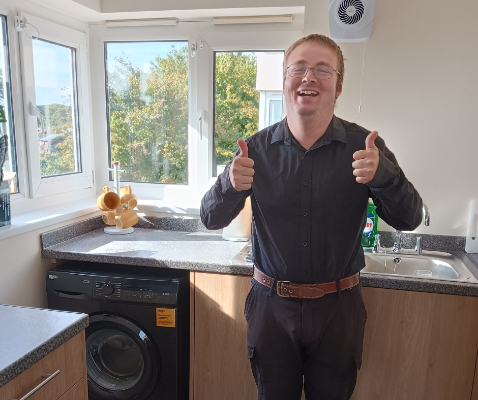 Andrew is a white man with ginger hair. He's standing in his new kitchen with two thumbs up and a beaming smile. He's so excited to be in his own place