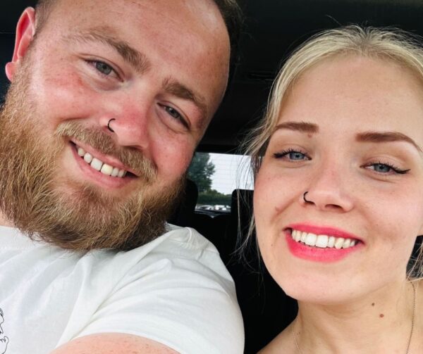 Chloe and her partner are taking a selfie together. They are both white, one male, one female. Chlose is blond, youthful looking, and wearing pink lipstick. She has her nose pierced and has blue eyes. Her partner also has his nose pierced and has a full beard and moustache. He has blue eyes too.