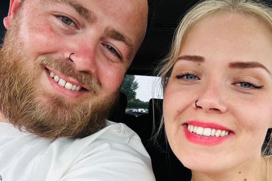 Chloe and her partner are taking a selfie together. They are both white, one male, one female. Chlose is blond, youthful looking, and wearing pink lipstick. She has her nose pierced and has blue eyes. Her partner also has his nose pierced and has a full beard and moustache. He has blue eyes too.