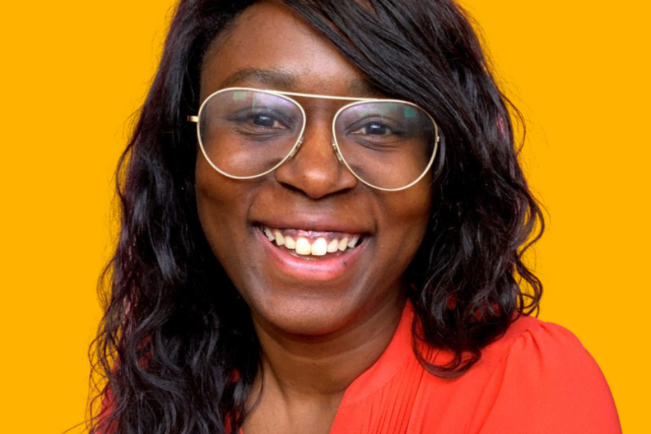 A headshot of Lisa Maynard-Atem who is a Black woman with long, dark brown wavy hair. She wears big, wide framed glasses that are very trendy. Her shoulder is just poking into the frame and she's wearing a read shirt. She looks really friendly and has a big smile on her face.