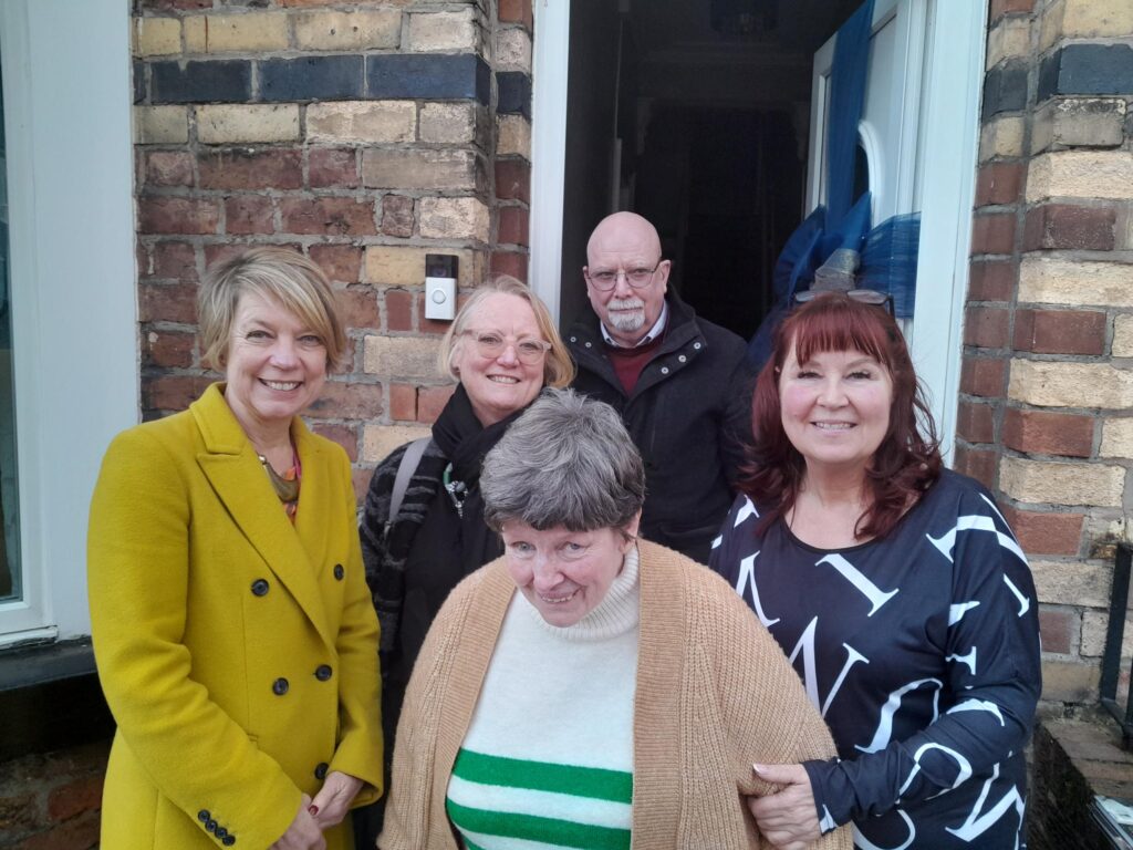 There's five people in the photo. They are all stood outside of the home of Jacqui an Sue's. Lesley is far left, white, blonde, average height and wears a mustard pea coat. She has short hair and is looking friendly - she's the former CEO of PSS. Julienne Meyer is next to Lesley and also blonde with short hair and wears a black winter coat. Sue is in front - she's an older lady with short grey hair, she wears a jumper with a cardigan over it and smiles brightly. Her Shared Lives carer, Jacqui is younger with dyed red hair, and a lovely smile - she holds Sue's arm to help keep her stable. She looks really warm and welcoming. Bob is at the back - he's the service manager. He's white, bald, and has a white goatee and glasses on as well as a dark coat and jumper.