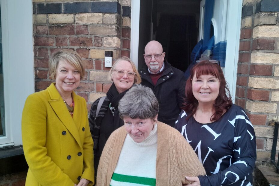 There's five people in the photo. They are all stood outside of the home of Jacqui an Sue's. Lesley is far left, white, blonde, average height and wears a mustard pea coat. She has short hair and is looking friendly - she's the former CEO of PSS. Julienne Meyer is next to Lesley and also blonde with short hair and wears a black winter coat. Sue is in front - she's an older lady with short grey hair, she wears a jumper with a cardigan over it and smiles brightly. Her Shared Lives carer, Jacqui is younger with dyed red hair, and a lovely smile - she holds Sue's arm to help keep her stable. She looks really warm and welcoming. Bob is at the back - he's the service manager. He's white, bald, and has a white goatee and glasses on as well as a dark coat and jumper.