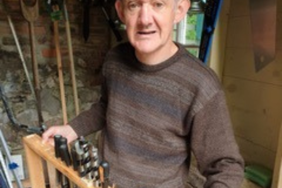 Trevor standing in his workshop, holding a wooden rack filled with neatly arranged woodworking tools. He is wearing a brown sweater, and the background shows various tools hanging on the wall, including spades and saws.