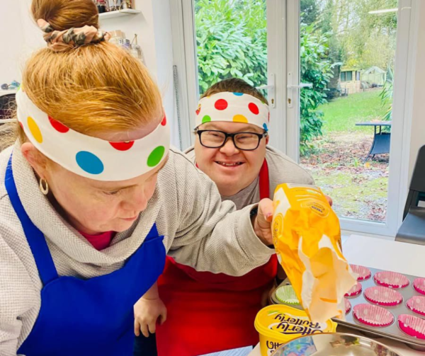 Victoria and someone who also uses Helena's day centre is cooking in the kitchen. They're wearing colourful polkadot headbands and aprons and Victoria is concentrating on weighing out flour. Her friend int he picture also has Down's Syndrome and smiling brightly at the camera. They're making cupcakes.