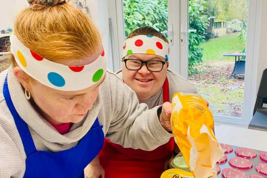 Victoria and someone who also uses Helena's day centre is cooking in the kitchen. They're wearing colourful polkadot headbands and aprons and Victoria is concentrating on weighing out flour. Her friend int he picture also has Down's Syndrome and smiling brightly at the camera. They're making cupcakes. Fostering adults can be as simple as baking a cake some days.