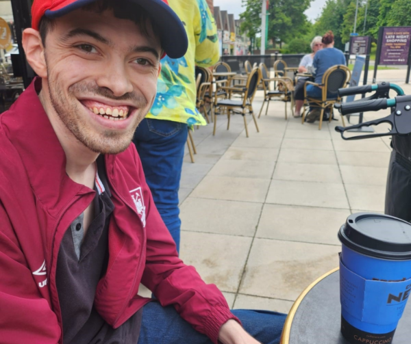 James, a white male in his 20s wears a red cap and a red jacket and jeans. He's outside a coffee shop enjoying a taekaway coffee and smiling brightly at the camera. He's clearly out and about in his community and loving it.