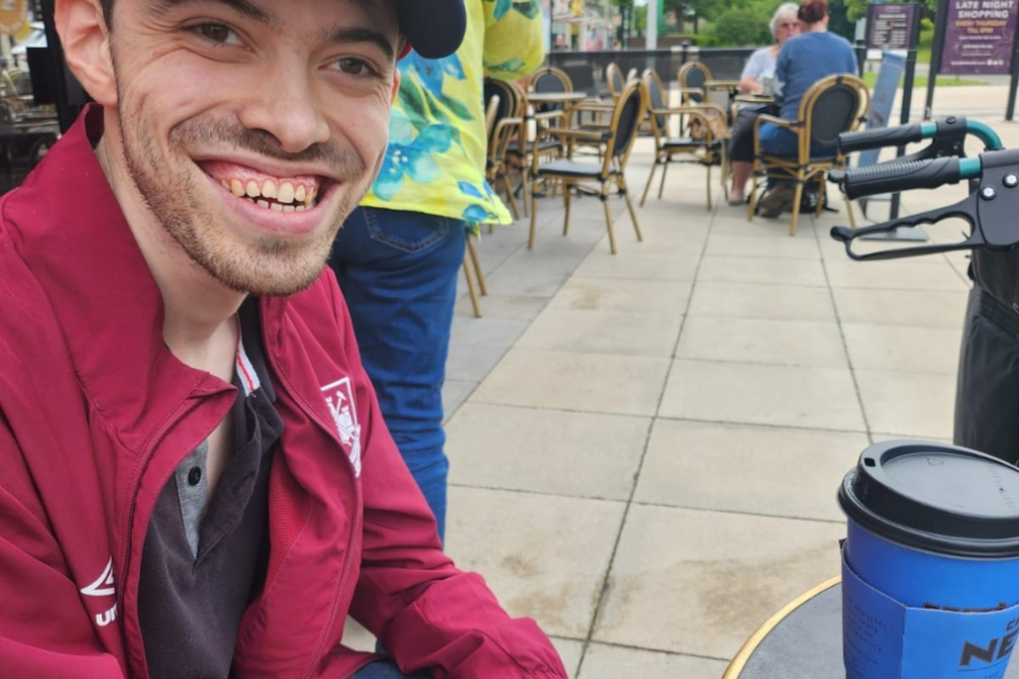 James, a white male in his 20s wears a red cap and a red jacket and jeans. He's outside a coffee shop enjoying a taekaway coffee and smiling brightly at the camera. He's clearly out and about in his community and loving it.