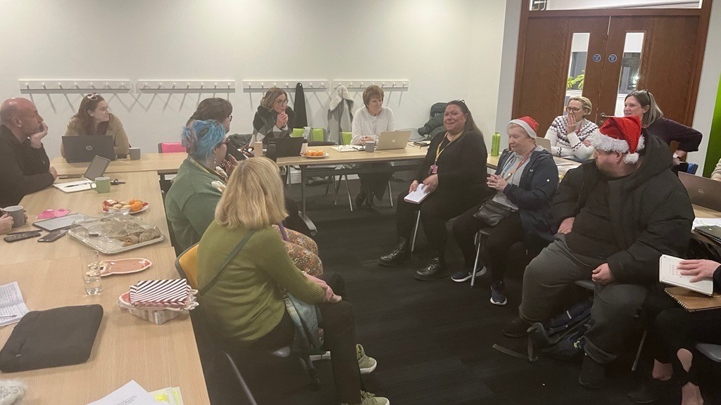 The picture shows a candid photo of the board meeting - it's not a stuffy board room look - the tables are packed with people of all different support needs as well as trustees. Everyone looks comfortable in their presence.