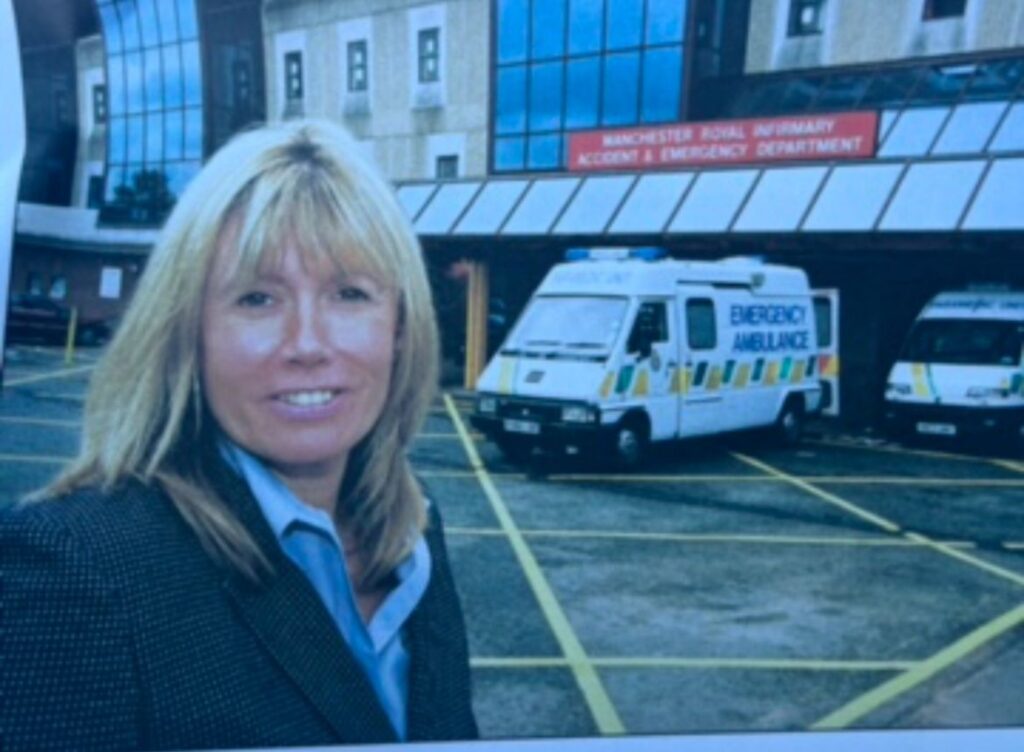 Janet is blonde and wearing a suit jacket, She is smiling at the camera in a friendly way. She's stood outside an A&E unit due to the nature of her work as a police officer back then.