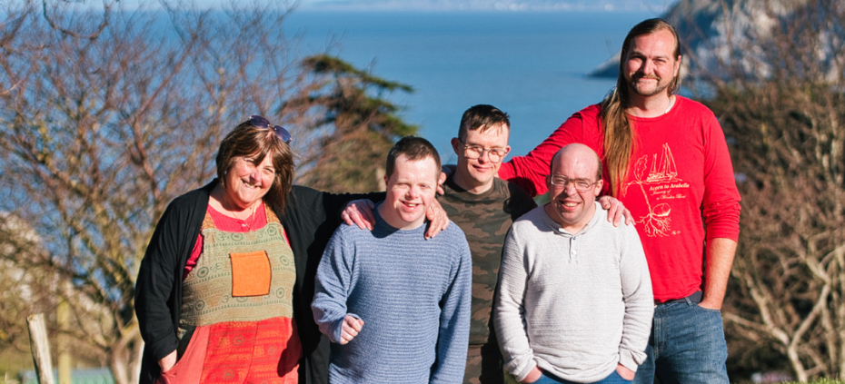 A Shared Lives family stand proudly together, smiling and happy - they're outdoors with Welsh countryside behind them. They look very comfortable together, just like family would.